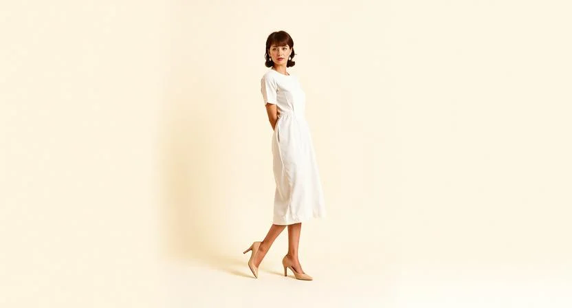 An elegantly dressed woman in a tailored ivory shift dress styled with kitten heels and pearl accessories in early 1960s fashion